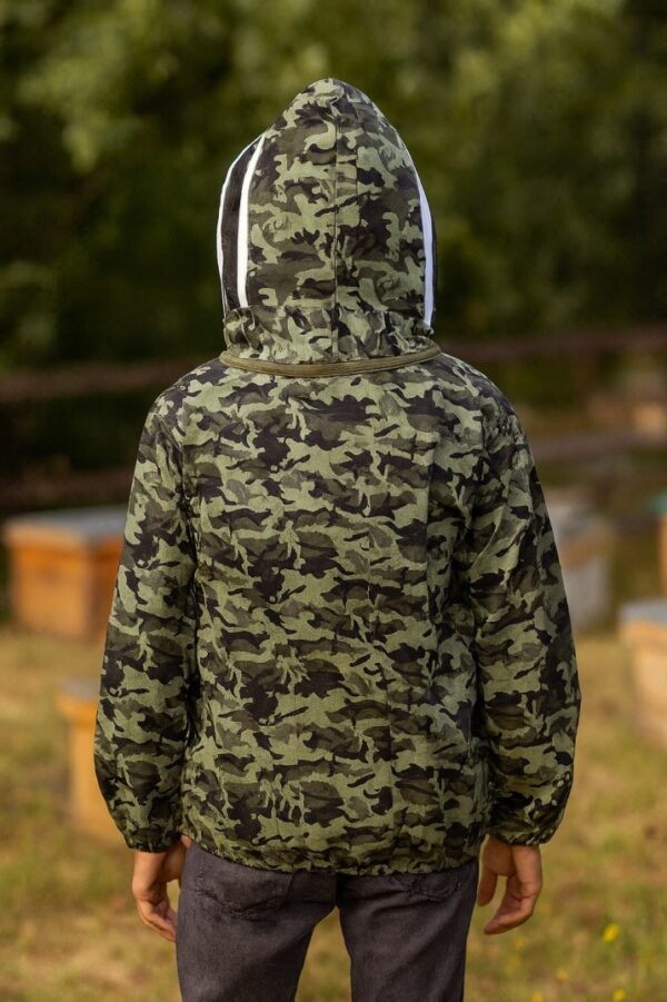 A man wearing a camouflage-patterned beekeeping suit with a white-trimmed mesh hood stands in front of wooden beehives. His face is faintly visible through the mesh veil, and he maintains a neutral expression. The background shows another person tending to a beehive, surrounded by grass and trees, creating an outdoor beekeeping scene.