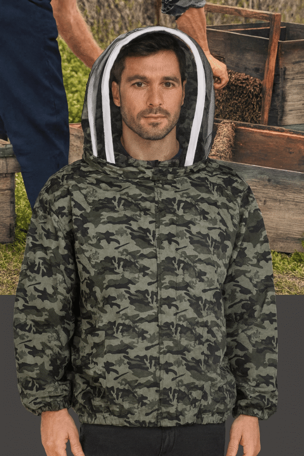 A man wearing a camouflage-patterned beekeeping suit with a white-trimmed mesh hood stands in front of wooden beehives. His face is faintly visible through the mesh veil, and he maintains a neutral expression. The background shows another person tending to a beehive, surrounded by grass and trees, creating an outdoor beekeeping scene.